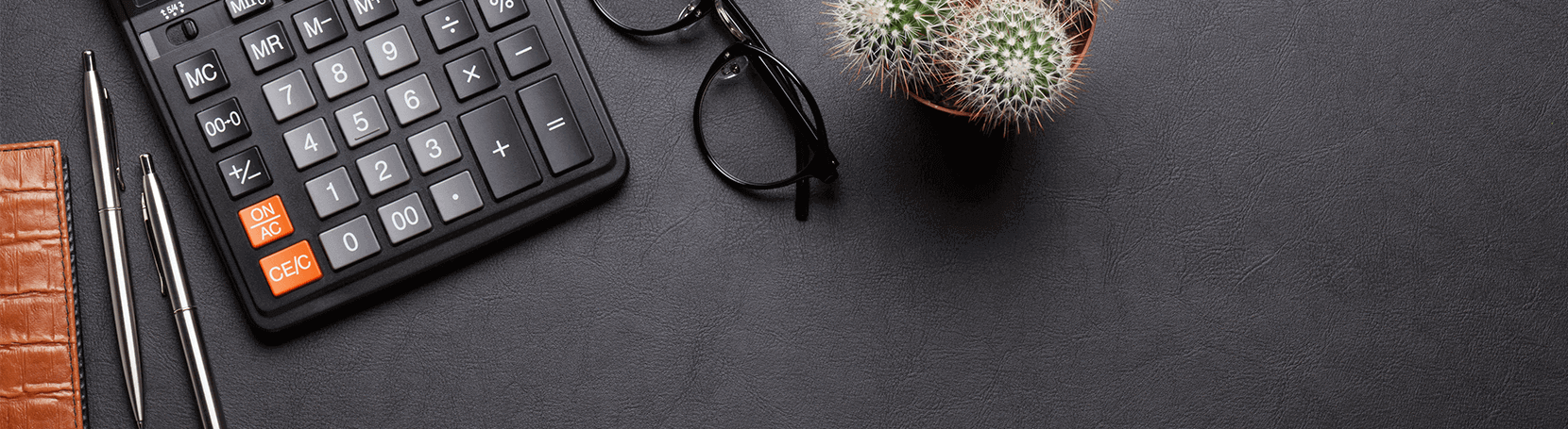 Calculator on a desk with a notebook, pens, glasses and a plant