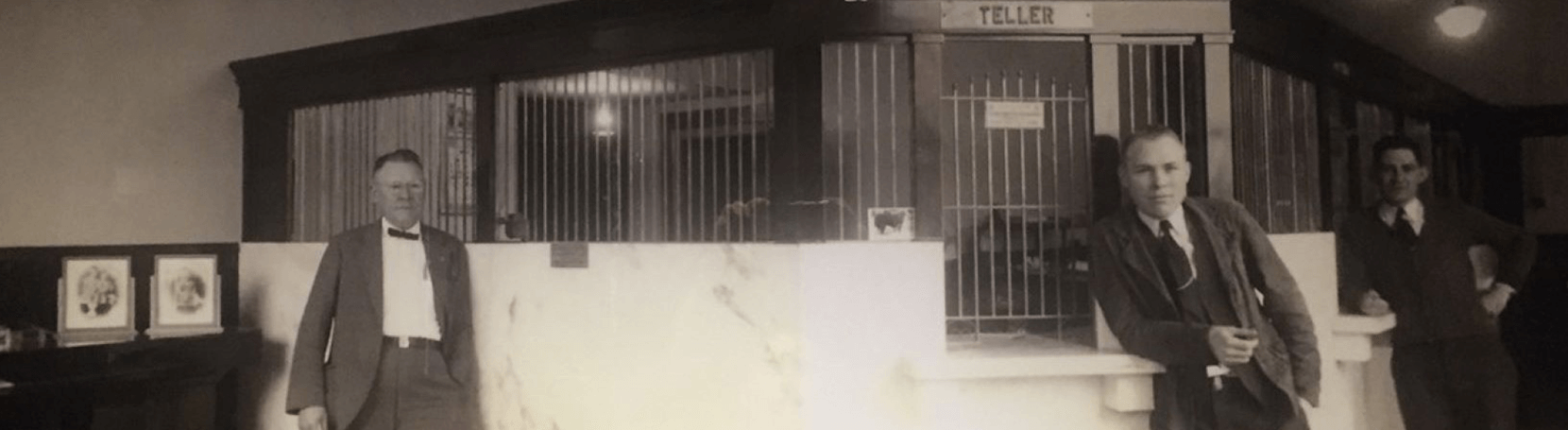 An old photo with three men standing in a bank next to a teller window.